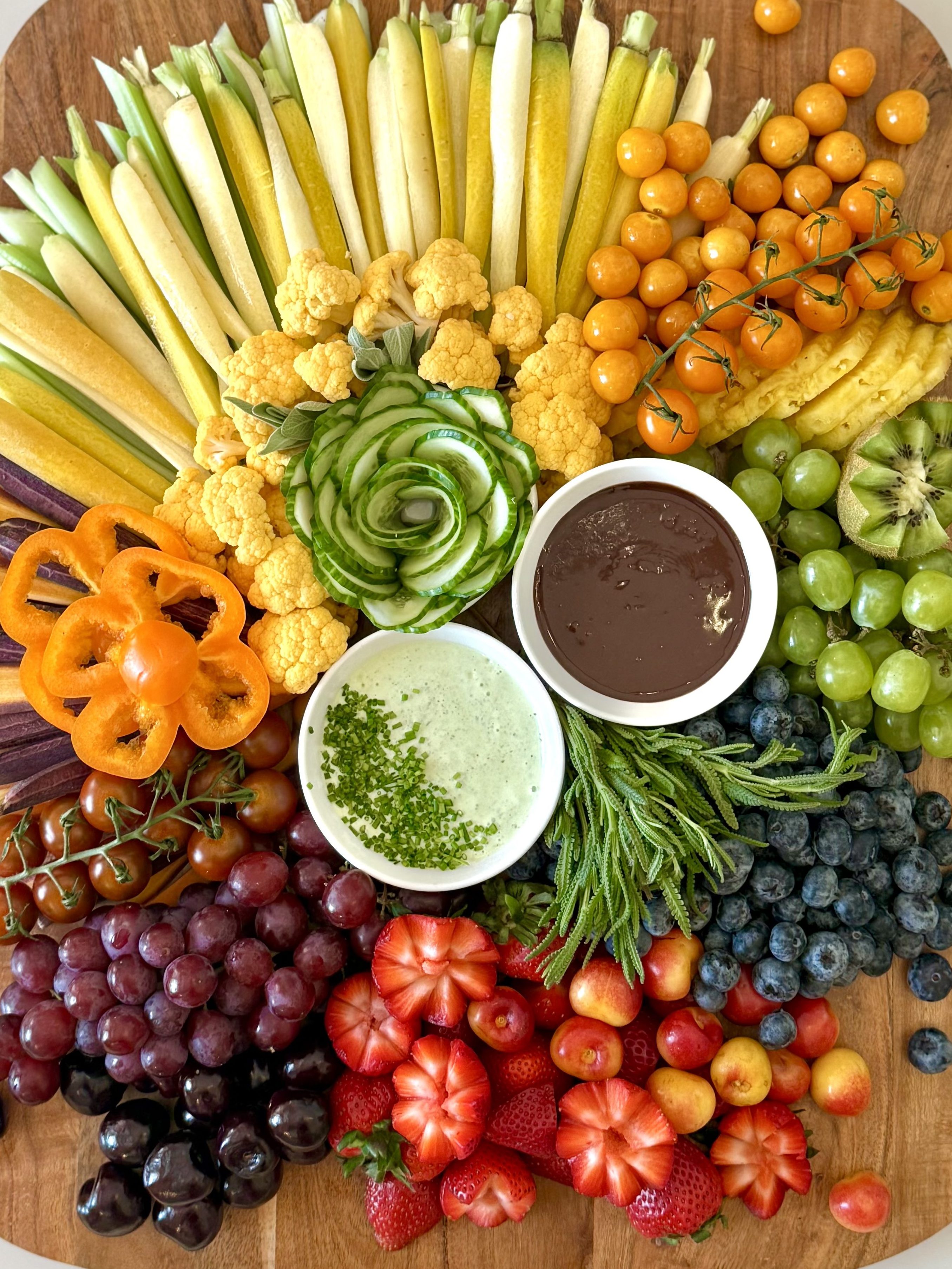 A rainbow-like board with sliced squash, cauliflower, tomatoes, bell peppers, grapes, strawberries, blueberries, and two dips—one chocolate and one green herb ranch.