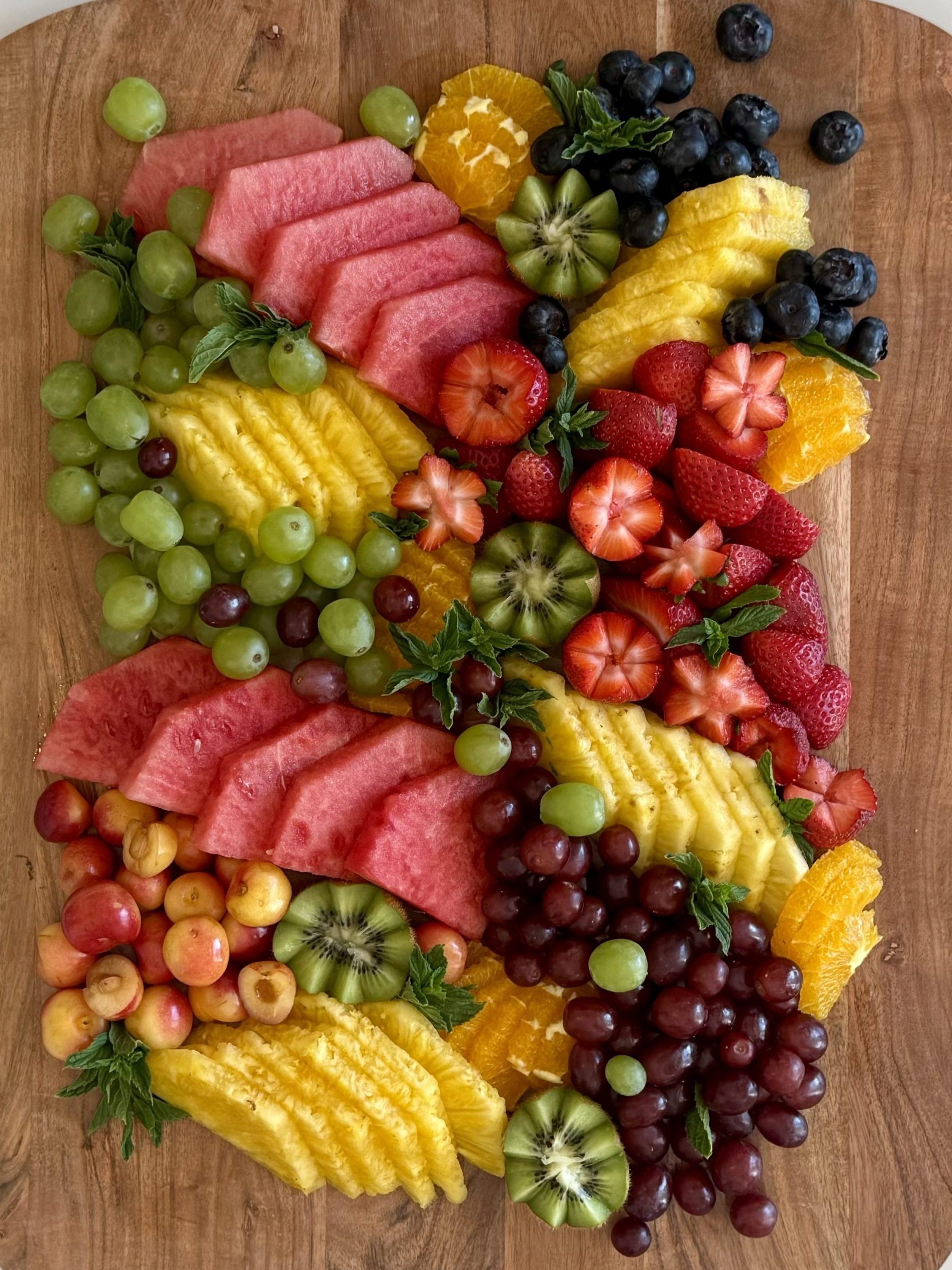 A fruit-only platter with sliced watermelon, pineapple, kiwi, green and red grapes, cherries, strawberries, figs, and orange slices, arranged in bold diagonal sections on a wooden board.